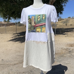 May include: A white short-sleeved dress featuring a graphic print of a city street scene with a yellow sign that reads CAFE. The lower portion of the dress is a cream-colored fabric with a subtle embroidered floral line art detail at the seam.