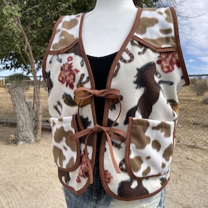 May include: A sleeveless fleece vest featuring a cream-colored base with a brown cow print, brown horse silhouettes, and red floral patterns. The vest has brown trim, two front pockets, and two brown ribbon ties at the center front closure.