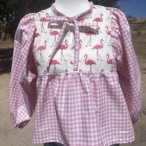 May include: A long-sleeved blouse featuring a white center panel with a pink flamingo print and a pink and white gingham pattern on the sleeves and lower body. The blouse has a round neckline with a tie closure and gathered sleeve cuffs.