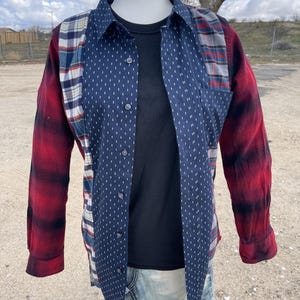 May include: A long-sleeve button-down shirt featuring a patchwork design. The center panel is dark blue with a white geometric pattern, flanked by sections of blue, white, and red plaid. The sleeves are red and black plaid with buttoned cuffs.