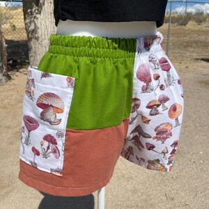 May include: Green, orange, and white shorts with a mushroom pattern. The shorts have an elastic waistband and pockets. The fabric is a soft material, perfect for casual wear.