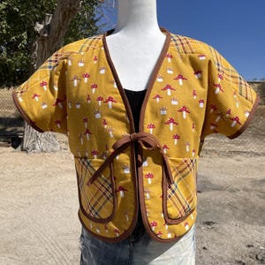 May include: A mustard yellow short-sleeved jacket featuring a repeating pattern of red and white mushrooms. The garment has brown trim, a front tie closure, and two patch pockets with a yellow, brown, and blue plaid fabric pattern.