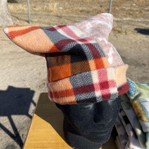 May include: A colorful fleece hat with a unique, pointed design. The hat features a patchwork of patterns in shades of orange, red, pink, and gray. The hat is displayed on a black mannequin head, showcasing its shape and style.