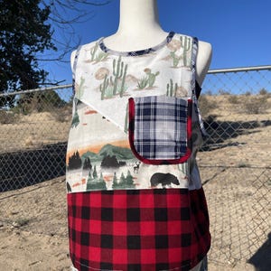 May include: A sleeveless top featuring a multi-panel design with a top section displaying a desert cactus print, a middle section showing a forest landscape with a bear, and a bottom section in red and black buffalo plaid with a matching plaid pocket.