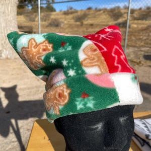 May include: A fleece beanie featuring a festive pattern of gingerbread men, holly berries, and white snowflakes on a green background, with a contrasting red section displaying white tree silhouettes and a wide white folded brim.