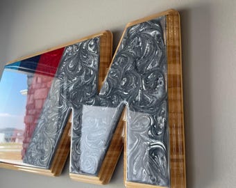 May include: A wooden wall art piece featuring the letter "M". The "M" is filled with a swirling silver and gray design. The left side of the "M" has a red, blue, and white stripe design. The piece is framed with light-colored wood.
