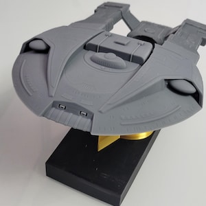 Könnte beinhalten: Ein graues 3D-gedrucktes Modell eines fiktiven Raumschiffs mit einem breiten, untertassenartigen Vorderteil mit komplexer Paneelstruktur, zwei nach hinten verlaufenden Triebwerkspylonen und einer zentralen rechteckigen Struktur.