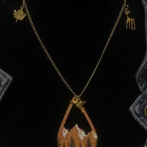 May include: A gold-colored necklace with a teardrop-shaped pendant featuring a mountain and forest scene. The necklace has a hummingbird charm and two additional charms: a maple leaf and a deer.