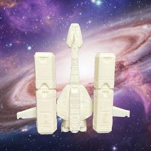 May include: A cream-colored plastic model of a spaceship featuring a central elongated fuselage with a pointed nose, flanked by two large rectangular engine blocks on either side, each with small wing-like protrusions extending outward.
