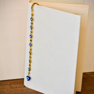 May include: A gold-colored bookmark with blue and white evil eye beads hangs from the top of a book. The bookmark is made of a chain with small, round gold-colored beads and blue and white evil eye beads. The book is open to a blank page.