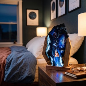 Peut inclure: Lampe décorative en bois avec un motif spatial. Elle présente un panneau en résine transparente avec des planètes, des étoiles et un vaisseau spatial. Le cadre en bois a une finition naturelle, et la lampe repose sur une base en bois. La lampe est sur une table de chevet en bois.