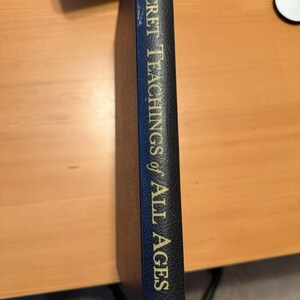 Puede incluir: El lomo de un libro de tapa dura negro con letras doradas que dicen The Secret Teachings of All Ages, con el logotipo de la Philosophical Research Society en la parte inferior. El libro tiene una superficie texturizada y est&aacute; colocado verticalmente.