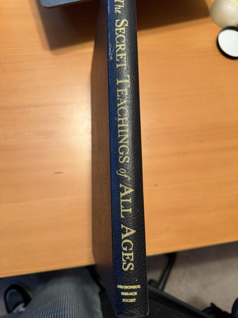 Puede incluir: El lomo de un libro de tapa dura negro con letras doradas que dicen The Secret Teachings of All Ages, con el logotipo de la Philosophical Research Society en la parte inferior. El libro tiene una superficie texturizada y est&aacute; colocado verticalmente.