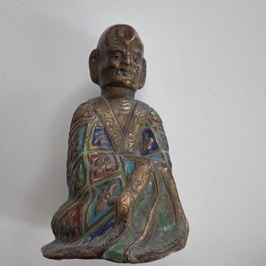 May include: A bronze-toned figurine of a seated figure with a bald head and prominent ears. The figure wears a robe featuring a grid pattern with colorful enamel inlays in shades of blue, green, and red, accented by intricate gold-toned scrollwork.