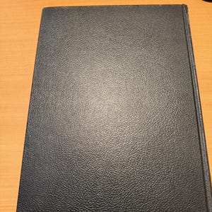 Puede incluir: Un libro de tapa dura rectangular con una cubierta de color azul marino oscuro que presenta una textura distintiva similar al cuero granulado. El libro est&aacute; colocado verticalmente, mostrando una superficie frontal limpia y s&oacute;lida con el lomo visible a la derecha.