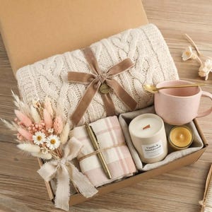Puede incluir: Una caja de regalo con una manta de punto trenzado color crema atada con una cinta de terciopelo marrón, una taza rosa con una cuchara dorada, una vela, un pequeño ramo de flores secas, un bolígrafo y una tela a cuadros rosa.