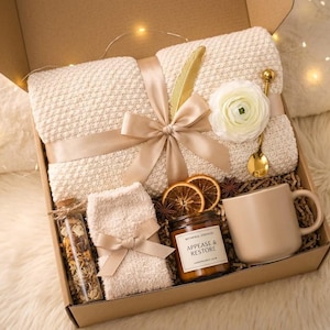 May include: A gift box filled with a cream-colored blanket, a mug, a candle, a small jar, a pair of socks, and decorative items. The blanket is tied with a ribbon. The candle has a label that reads "APPEASE & RESTORE".
