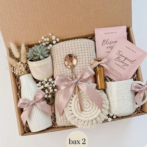May include: A gift box containing self-care products. It features a white mug, a small succulent in a pot, a vase, a waffle-knit blanket, a spoon, tea sachets, a macrame coaster, and a small bottle. All items are tied with pink ribbons.