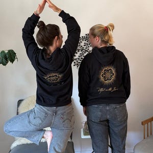 Op de afbeelding: Twee mensen dragen zwarte hoodies met gouden bloemmotieven en de tekst "Made with Love". De hoodies hebben trekkoorden en een voorzak. Ze worden gecombineerd met jeans. Eén persoon balanceert op één been.