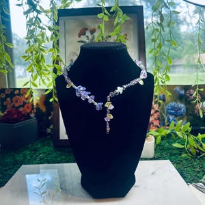 May include: A necklace featuring a series of translucent purple and yellow flower-shaped beads interspersed with small clear crystals and silver-toned accents. The design includes a central dangling element with floral beads and a delicate chain.
