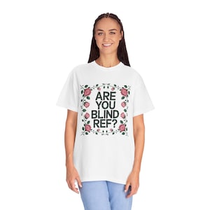 May include: White t-shirt with the text "ARE YOU BLIND REF?" in black, framed by a floral design with pink roses and green leaves. The shirt is a casual, comfortable style.