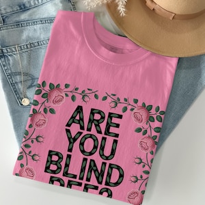 May include: A pink t-shirt with the text "ARE YOU BLIND DEER" in black letters, framed by a floral design of pink roses and green leaves. The shirt is laid on top of blue jeans and a tan hat.