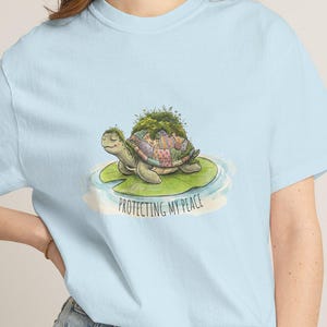 Peut inclure: T-shirt bleu clair avec un dessin de tortue de dessin animé. La tortue a une carapace recouverte de mousse et est posée sur une feuille de nénuphar, avec le texte "PROTECTING MY PEACE" en dessous. Le t-shirt est en tissu doux.
