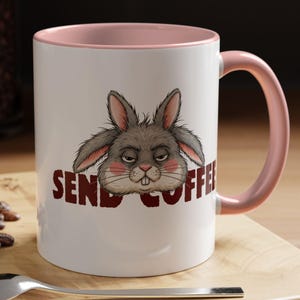 Könnte beinhalten: Weiße Keramik-Tasse mit rosa Innenraum und Henkel, mit einem mürrischen Cartoon-Kaninchen und den roten Worten "SEND COFFEE". Ein Löffel und ein Gebäck befinden sich im Vordergrund.