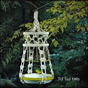 Może przedstawiać: Biały karmnik dla ptaków wykonany z makramu z żółtą i białą miską wiszącą na gałęzi drzewa. Karmnik jest wykonany z zawiązanej liny i ma ozdobny wzór. Tekst "Tick Tock Knits" jest widoczny w prawym dolnym rogu obrazu.