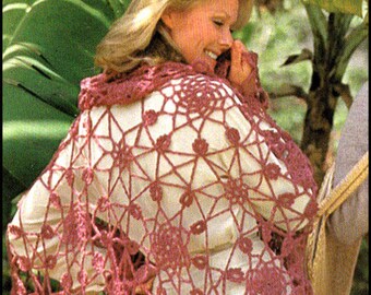 Puede incluir: Un chal de ganchillo rosa con un delicado patrón floral y flecos. El chal lo lleva una persona con el pelo rubio y está drapeado sobre un vestido blanco.