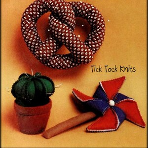 Puede incluir: Un pretzel de tela marrón y blanco con alfileres, un cactus verde en una maceta marrón y una veleta roja, blanca y azul con un palo de madera. El texto "Tick Tock Knits" está impreso en el fondo.