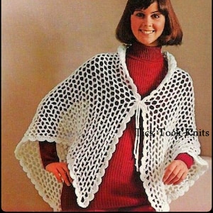 May include: White crochet shawl with a scalloped edge and a tie closure. The shawl is made with a delicate openwork pattern. The model is wearing a red turtleneck sweater under the shawl.
