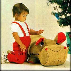 Peut inclure: Un jeune enfant portant une salopette rouge et une chemise blanche est agenouillé à côté d'un grand hippopotame en peluche brun tricoté. Le jouet a de grands yeux noirs et un nez rouge. L'enfant regarde le jouet avec un sourire.