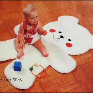 May include: A white knitted bear rug with a red nose and red cheeks. A baby is sitting on the rug, playing with toys. The rug is on a wooden floor. Tick Tock Knits.