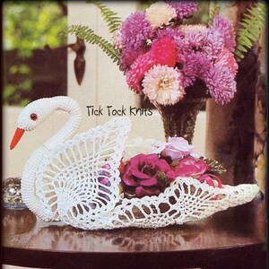 Può includere: Un cesto a forma di cigno bianco lavorato a uncinetto con un motivo floreale. Il cesto è pieno di fiori rosa e viola. Il testo "Tick Tock Knits" è visibile nell'immagine.