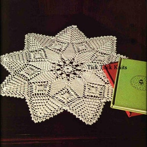 May include: A white crocheted doily with a starburst design. The doily is on a dark brown surface with two books, one red and one green, with the text "Tick Tock Knits" on the red book.