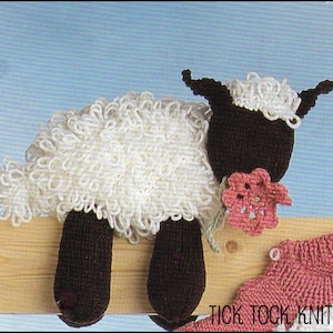 May include: A white knitted sheep with black legs and a pink flower. The sheep is sitting on a wooden surface. The text "Tick Tock Knits" is visible in the lower right corner of the image.