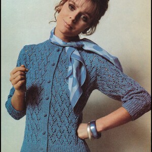 May include: A blue knitted cardigan with a button-up front and a diamond-shaped pattern. The cardigan is worn over a light blue skirt and a blue and white patterned scarf.