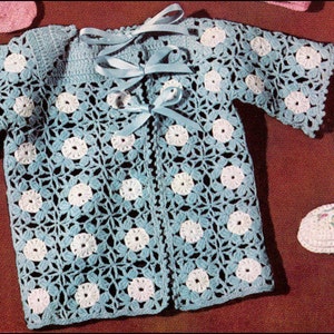May include: A crocheted baby cardigan with a blue and white floral pattern. The cardigan has a tie closure and short sleeves. The image also shows a pair of crocheted baby shoes with pink bows.