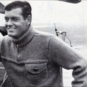 Op de afbeelding: Een zwart-witfoto van een man die een grijze gebreide trui draagt met een rits en een klepzak. De trui heeft een geribbelde kraag en manchetten. De man staat voor een wazige achtergrond.