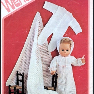 Puede incluir: Un conjunto de muñeca de bebé de punto blanco con un gorro a juego y una manta de ganchillo blanca. La muñeca está sentada en una pequeña silla de madera. El patrón se llama "Tick Tock Knits" y el número es 1813 DK.