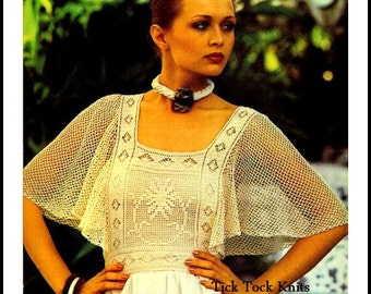 May include: A white crochet top with a square neckline and a delicate floral pattern. The top has a wide, flowing cape-like sleeve. The model is wearing a white dress and a brown and white necklace.