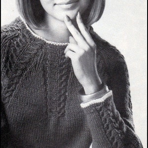 May include: A black and white photo of a woman wearing a knitted sweater with a cable knit pattern. The sweater has a white trim around the neckline and cuffs. The woman is sitting with her hand on her chin and looking at the camera.