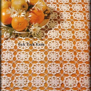 May include: White crocheted tablecloth with a repeating geometric pattern. The tablecloth is laid on a wooden surface and has a bowl of fruit and flowers on top. The text "Tick Tock Knits" is visible in the center of the tablecloth.