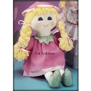 May include: A handmade doll with blonde yarn hair in pigtails, wearing a pink dress with green trim and a white hat. The doll is sitting on a blue background with the text "Tick Tock Knits" visible.