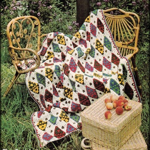 Op de afbeelding: Een kleurrijk gehaakt deken met een ruitpatroon in tinten rood, groen, geel en blauw. Het deken is gedrapeerd over een rieten stoel en een rieten mand gevuld met perziken.