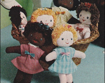 May include: A group of six handmade dolls in a wicker basket. The dolls are dressed in various patterns and colors, including red, white, blue, and plaid. The dolls have yarn hair and button eyes.