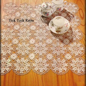 以下が含まれることがあります： 繊細な花柄の白いクロシェのドイリー。ドイリーは木製の表面に置かれ、3つの磁器製のティーカップとソーサーが置かれています。画像には「Tick Tock Knits」というテキストが表示されています。