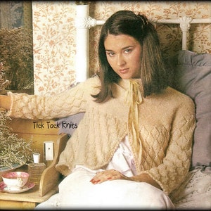 May include: A woman wearing a cream-colored knitted cardigan with a delicate lace pattern. The cardigan has a shawl collar and a tie at the neck. The woman is sitting on a bed with a floral patterned wallpaper.