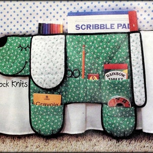 May include: Green fabric dog-shaped organizer with pockets for storing crayons, pencils, scissors, and other supplies. The organizer has a white fabric lining and black trim. The text "Tick Tock Knits" is printed on the organizer. The organizer is hanging on a white bed skirt with blue polka dots.